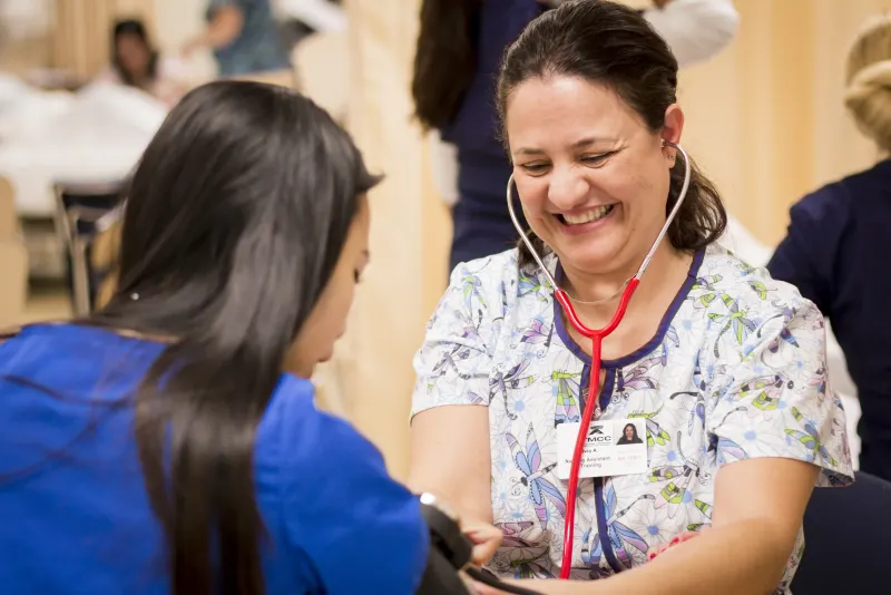 Certified nursing assistant student taking a classmate's blood pressure with a stethoscope in a training lab