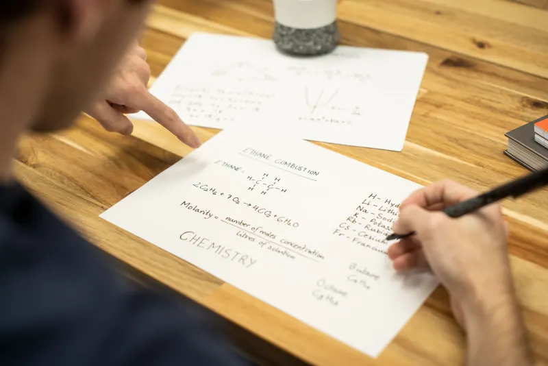 Student working through handwritten chemistry notes at a wooden desk while a tutor points to a formula