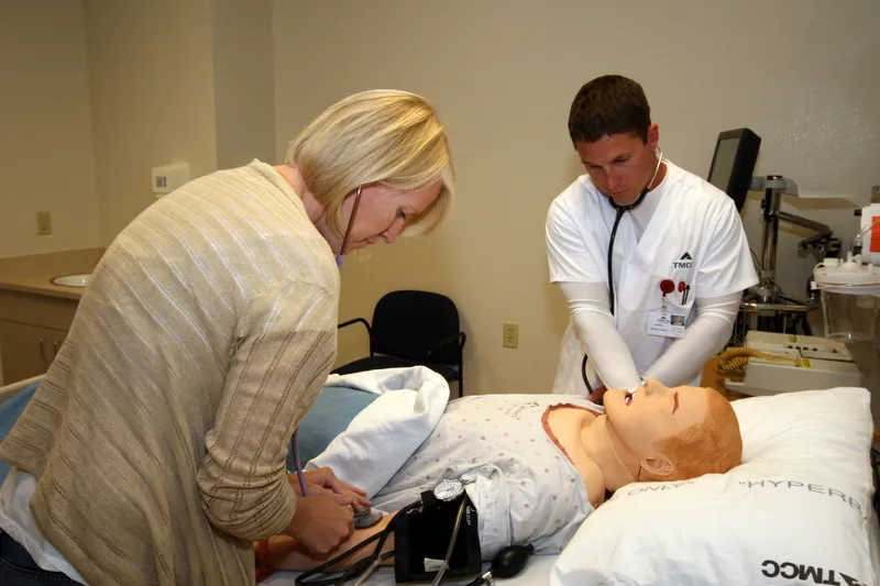 Nursing student checking a patient simulator mannequin with a stethoscope while an instructor checks its blood pressure in a college nursing lab